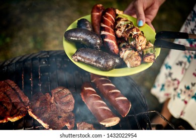 Juicy Sausages Straight From Grill On Green Plate Against The Background Of Hot Barbecue On Summer Day