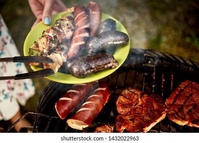 Juicy Sausages Straight From Grill On Green Plate Against The Background Of Hot Barbecue On Summer Day