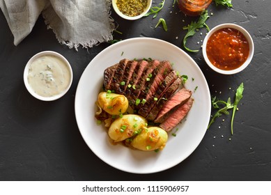 Juicy steak medium rare beef with baked potatoes on white plate over black stone table. Top view, flat lay