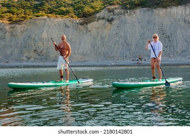 July 5, 2022. Dalaman, Turkey. Older Couple On Paddle Board On Quiet Sea With Rocky Coastline And Morning Sunlight. Active Lifestyle For Aged Man And Woman