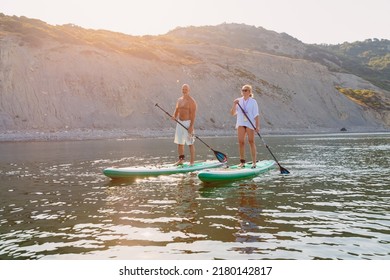 July 5, 2022. Dalaman, Turkey. Older Couple On Paddle Board On Quiet Sea With Rocky Coastline And Morning Sunlight. Active Lifestyle For Aged Man And Woman