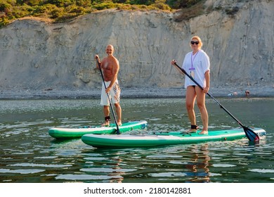 July 5, 2022. Dalaman, Turkey. Older Couple On Paddle Board On Quiet Sea With Rocky Coastline And Morning Sunlight. Active Lifestyle For Aged Man And Woman