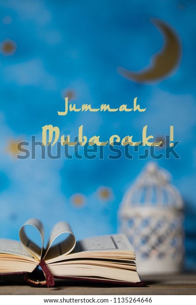 Jummah\
Mubarak! Happy Friday! Sacred book of Quran on a wooden surface.\
Blue night sky and candle holder in the\
background
