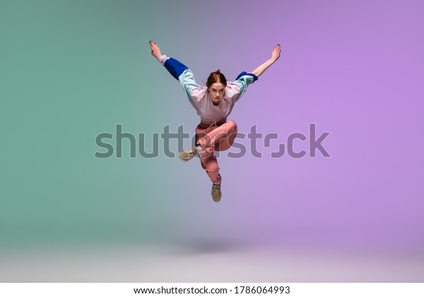 In jump. Beautiful girl dancing hip-hop in\
stylish clothes on colorful gradient background at dance hall in\
neon light. Youth culture, movement, style and fashion, action.\
Fashionable bright portrait.