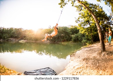 Jump Into The Water. A Man Is Resting On The Nature. A Swing From A Rope And A Stick. Active Recreation In Nature. Friends Have Fun On The Lake. Fisheye Lens.