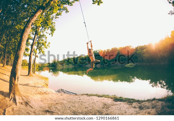 Jump into the water. A man\
is resting on the lake. A swing from a rope and a stick. Active\
recreation in nature. Summer fun. A man is riding a swing. Fisheye\
lens.