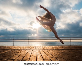 Jumping ballerina in beige dress and pointe on embankment above ocean or sea beach at sunrise.Beautiful blonde woman with long hair practicing stretching and classic exercises.Epic jump.
