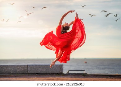 Jumping ballerina in a red flying skirt and leotard on ocean embankment or sea beach surrounded by seagulls in the sky.