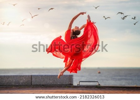 Jumping ballerina in a red flying skirt and leotard on ocean embankment or sea beach surrounded by seagulls in the sky.