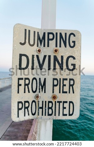 Jumping Diving From Pier Prohibited written on a sign board in Destin Florida. The ocean, horizon and sky can be seen in the blurry background of the warning signage.