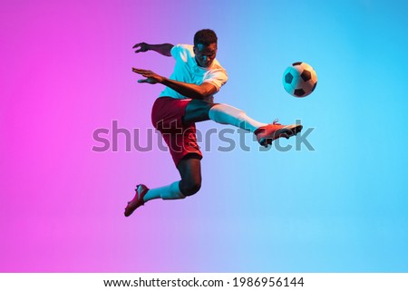 Jumping, flying. One young African man, professional soccer football player training isolated on gradient blue pink background in neon light. Concept of action, energy, sport. Copy space for ad