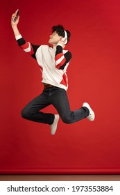 Jumping High, Listening To Music. Asian Man's Portrait Isolated On Red Studio Background With Copyspace For Ad. Handsome Man With Devices. Concept Of Human Emotions, Facial Expression, Youth Culture.