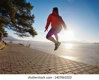 Jumping Silhouette Against Low Sun. Workout At The Lake Beach. Winter Morning Excercise, Ice Cover Lake And Create Nice Reflection