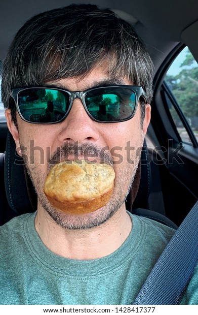 June 16, 2019. A man is eating a banana muffin
while driving a car, holding the muffin in his mouth without using
his hands. He’s wearing sunglasses, has his seatbelt fastened.
Eating while driving.