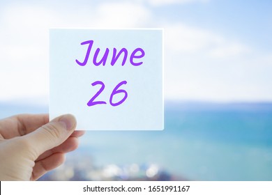 June 26th. Hand Holding Sticker With Text June 26 On The Blurred Background Of The Sea And Sky. Copy Space For Text. Month In Calendar Concept