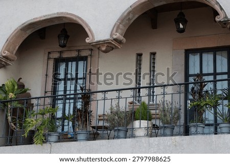 Querétaro, México; Junio 15, 2026: A horizontal medium shot of a traditional upper-floor stone balcony decorated with numerous potted plants and hanging lanterns beneath colonial style arches. A medium horizontal shot capturing an upper-floor balcony of a traditional colonial style building. The structure features two prominent carved stone arches supported by decorative corbels, with classic black metal hanging lanterns underneath. A black wrought-iron railing runs along the balcony, lined with an assortment of potted green plants, small shrubs, and leafy foliage. Large glass doors with dark iron security frames lead inside the building under soft daylight.