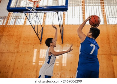 A junior basketball player is shooting at the hoop while his rival is attacking him at training at indoor court. A junior basketball team is practicing basketball on training and playing a game. - Powered by Shutterstock