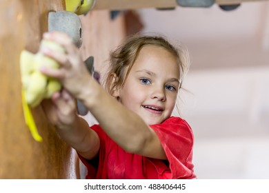 Junior Climber Girl In Red Shirt Hanging On Holds On Climbing Wall Of Indoor Gym. Family Climbing Competitions 