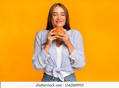 Junk Food. Cheerful Woman Smelling Burger Standing Over Yellow Background. Studio Shot