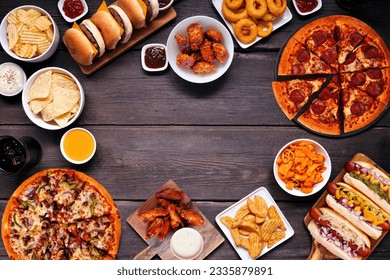 Junk food frame. Pizza, hamburgers, chicken wings, hot dogs and salty snacks. Top view over a dark wood banner background with copy space. - Powered by Shutterstock