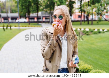 Junk food. Happy delighted woman in casual clothes eating donut with expression of big pleasure, temptation to bite doughnut, appetizing bakery. Sweet tooth,indulgence, gluttony, unhealthy lifestyle.