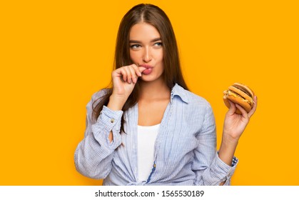 Junk Food Lover. Hungry Girl Holding Burger Licking Fingers Standing Over Yellow Studio Background.