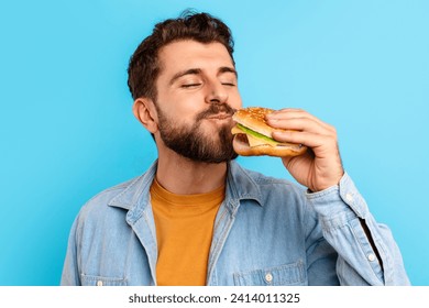 Junk Food Lover. Portrait Of Cheerful Guy Posing With Burger, Eating Big Tasty Sandwich On Blue Studio Background, Closeup Shot. Bad Nutrition And Overeating Habit, Fast Food Concept - Powered by Shutterstock