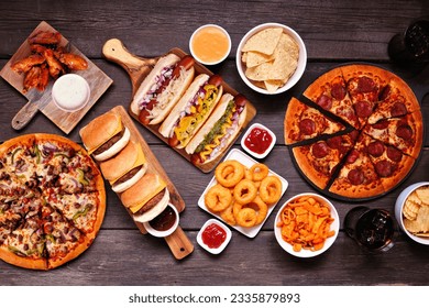 Junk food table scene. Pizza, hamburgers, chicken wings, hot dogs and salty snacks. Above view over a dark wood background. - Powered by Shutterstock