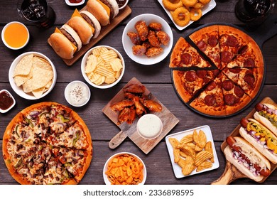 Junk food table scene. Pizza, hamburgers, hot dogs, chicken wings and salty snacks. Overhead view over a dark wood background. - Powered by Shutterstock