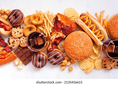 Junk food table scene scattered over a white marble background. Collection of take out and fast foods. Pizza, hamburgers, french fries, chips, hot dogs, sweets. Top view.