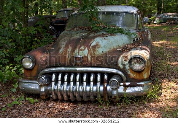Junk yard vehicles showing old rusted car in\
overgrown weedy area