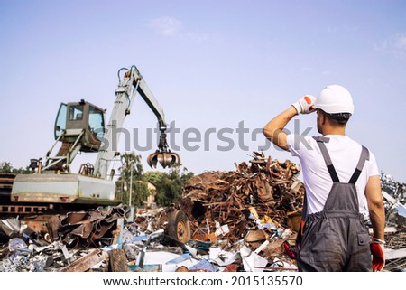 Junkyard worker supervisor looking at industrial crane machine lifting metal parts for recycling.