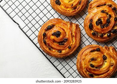 Just baked pain aux raisins on cooking wire rack. Buns are also called escargot or pain russe, is a spiral pastry with custard cream and raisin. Directly above, white table surface.