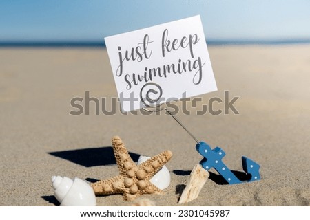 JUST KEEP SWIMMING text on paper greeting card in anchor paper holder and starfish seashell summer vacation decor. Sandy beach sun coast. Holiday concept postcard. Getting away Travel