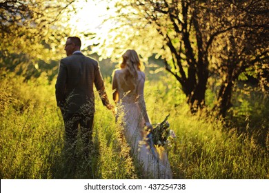 Just Married Loving Hipster Couple In Wedding Dress And Suit On Green Field In A Forest At Sunset. Happy Bride And Groom Walking Running And Dancing In The Summer Meadow. Romantic Married Young Family