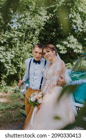 Just married wedding couple is standing near the retro vintage car in the park. Summer sunny day in forest. bride in elegant white dress with bouquet and elegant groom in love hugging. Bride and groom
