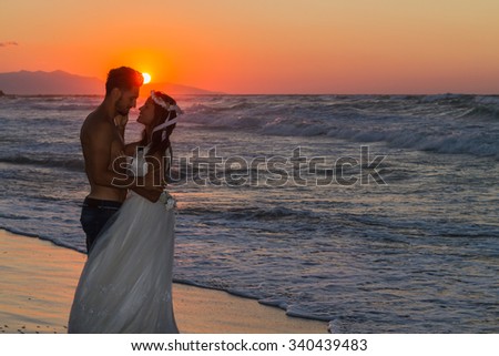 Just married young couple at the beach, enjoying the hazy dusk, wearing a wedding dress and shorts, walking barefoot, getting wet, teasing and kissing one another.