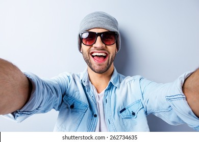 Just Me And No One Else. Top View Of Handsome Young Man In Hat And Sunglasses Making Selfie And Smiling While Standing Against Grey Background