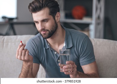 Just One Pill Can Help. Handsome Young Man Holding A Glass Of Water And Looking At A Pill In His Hand While Sitting On The Couch At Home