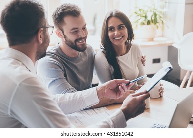 Just put your signature here! Confident young man in shirt and tie holding some document and pointing it while sitting together with cheerful young couple at the desk in office  - Shutterstock ID 431851042