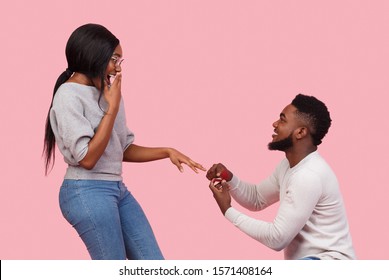 Just say yes. Romantic afro guy making proposal to his amazed woman, staying on knee with ring over pink background, copy space