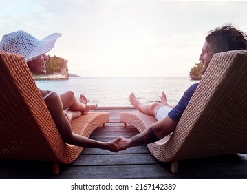 Just the two of us. Rearview shot of a cheerful young couple holding hands while relaxing on deck chairs and looking out at the ocean outside during the day.