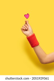 K Pop Concept. A Girl Hand With Red Sport Cotton Sweat Bands Showing Fingers Heart Gesture. Red Glitter Heart Above. Optimistic Yellow In Background.