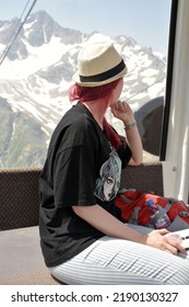 Kabardino-Balkaria, Russia, July 18, 2022 - A Girl In A Hat Sits In A Funicular Cabin And Looks At The Beautiful Snowy Mountain Slopes. Mountains Of The Caucasus.