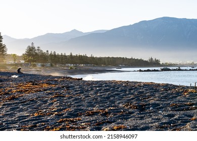 Kaikoura New Zealand - April 11 2022; View Across Kaikoura Beach At Sunset With Couple Sitting On Stones In Hazy Light.