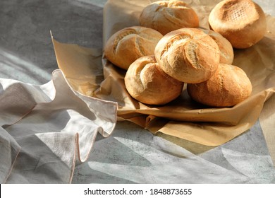 Kaiser or Vienna rolls in bread basket with towel. Table covered with beige linen tablecloth. Sun light with long shadows.