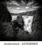 Kakabeka Falls, the second largest waterfall in Ontario (behind only Niagara Falls) plunges down a rocky cliff, as seen from a lookout point. Black and white.