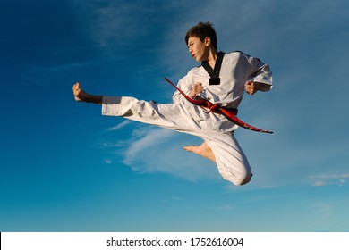 Karate boy kicks in the air and flies over the blue background of the sky. Practicing Taekwondo in nature. Black martial arts belt