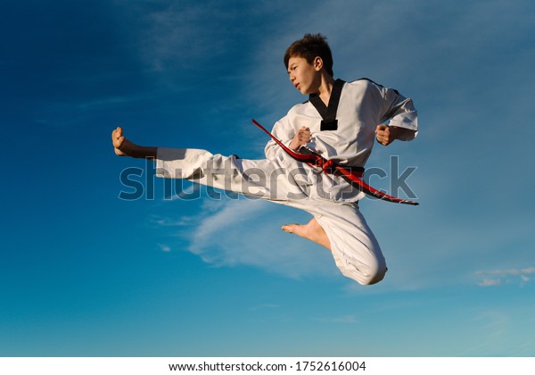 Karate boy kicks in the air and flies over the blue\
background of the sky. Practicing Taekwondo in nature. Black\
martial arts belt