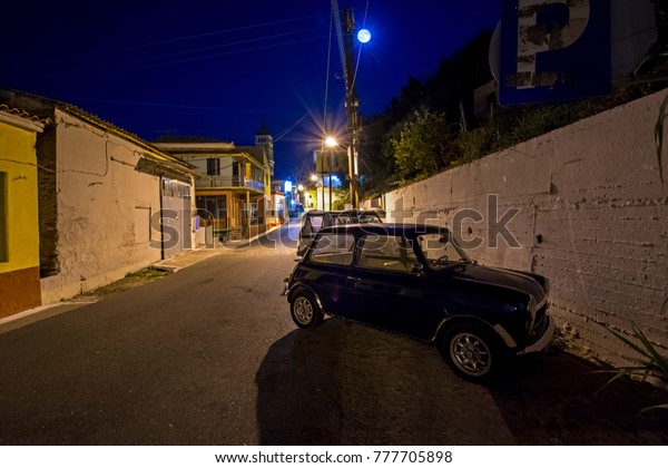Karousades, Corfu, Greece - August 31, 2017: night\
cityscape of old mediterranean village with navy blue Austin Mini\
Cooper 1000  on narrow street with antique houses under the full\
moon 